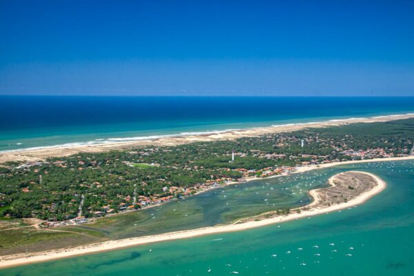 Vue aérienne du Bassin d'Arcachon avec le phare du Cap Ferret et l'océan en arrière-plan. La mer, d'un bleu et vert éclatant, entoure un banc de sable, tandis que des bateaux au mouillage complètent ce paysage marin paisible, Gerald Photography auteur photographe